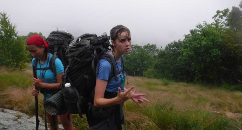 Two people wearing backpacks stand in a grassy meadow surrounded by trees. One of them gives an "I love you" hand gesture to the camera. 